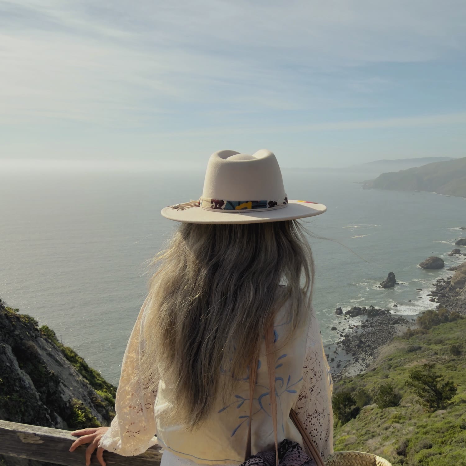A woman in a wide-brimmed hat stands at a wooden railing at Muir Beach Overlook in Marin, her back to the camera, looking out over the Pacific.
